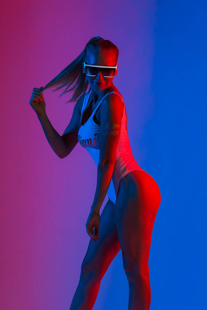 Portrait of positive fashion young girl in a white swimsuit and stylish sunglasses in red and blue and purple neon light. Studio shot. - 写真・画像