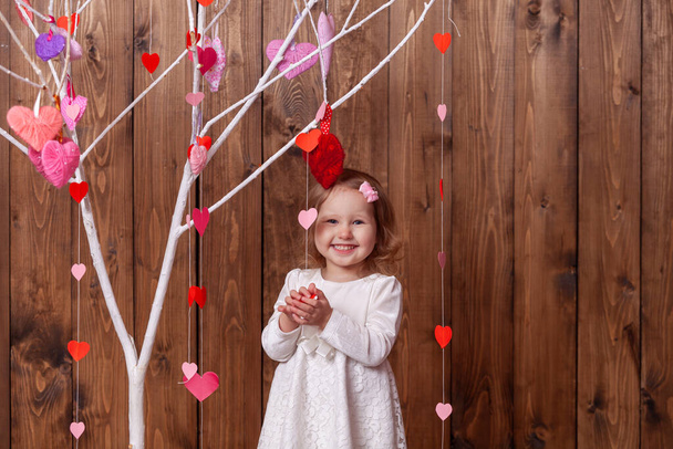 happy little girl on a beautiful background for a photo shoot for Valentine's Day looks out from behind a tree with red and pink hearts. A cheerful child celebrates Valentine's Day - Fotografie, Obrázek