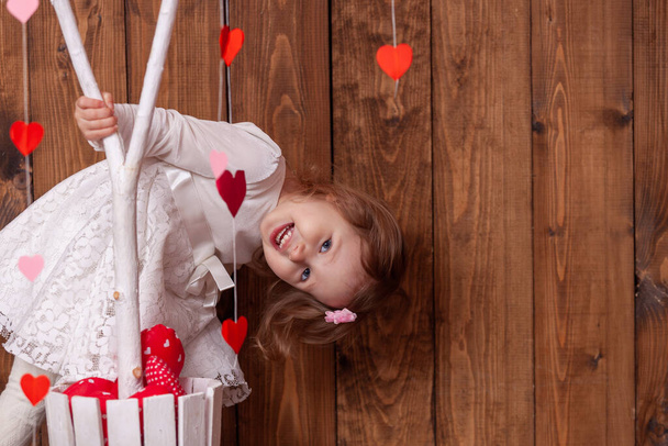 happy little girl on a beautiful background for a photo shoot for Valentine's Day looks out from behind a tree with red and pink hearts. A cheerful child celebrates Valentine's Day - Foto, Imagem