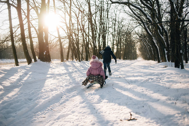 Happy boy tire une fille sur un traîneau, marchant à l'extérieur par une journée d'hiver glacée. Merveilleux soleil du soir. - Photo, image