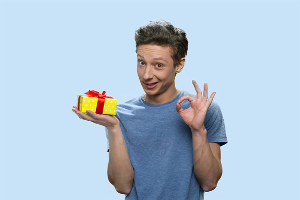 Smiling boy holding a gift box and showing ok gesture. - Foto, Bild