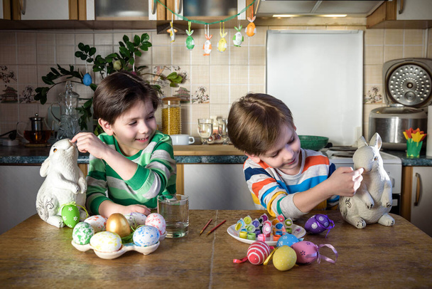 Dva malí kluci zbarvují vajíčka na velikonoční prázdniny v domácí kuchyni, uvnitř. Bratři sourozenci se baví a slaví hostinu. - Fotografie, Obrázek