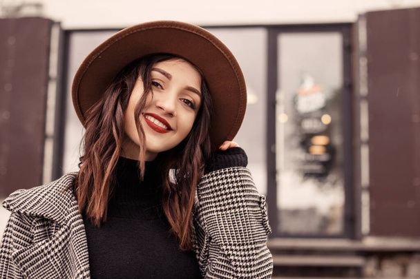 Close up shot charming woman standing outside near street cafe restaurant wear brown hat gray coat poloneck black sweater, looking at the camera. Girl spend time autumn spring, cold weather fresh air - Foto, Imagem
