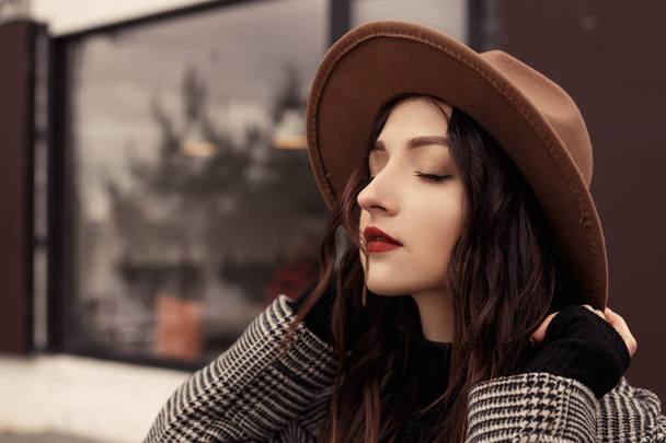 Close up shot urocza kobieta stojąca na zewnątrz w pobliżu restauracji kawiarnia nosić brązowy kapelusz szary płaszcz poloneck czarny sweter, odwracając wzrok. Dziewczyna spędzają czas jesienią lub wiosną, zimna pogoda świeże powietrze. - Zdjęcie, obraz
