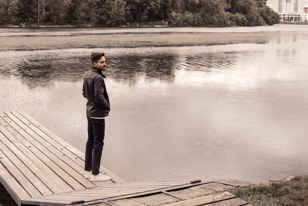 深刻な思慮深い男の完全な長さの肖像画は、川の湖の霧の雨の天気の近くの木製の桟橋に立って、離れて見て。ドレスは黒革暖かいジャケットを着用してください。濁った水が自然を流れる - 写真・画像