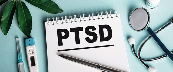 In a white notebook on a blue background, near a sheet of shefflers, a stethoscope, a syringe and an electronic thermometer, the word PTSD is written. Medical concept - Photo, image