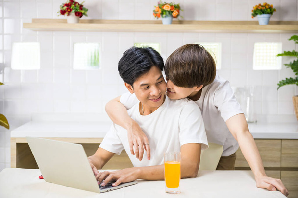 LGBT-Männer arbeiten am Laptop, während seine Partnerin ihn im Wohnzimmer von hinten umarmt und küsst. Süße männliche homosexuelle Paar verbringen Zeit zu Hause. Sie lachen und schauen einander an. Gleichgeschlechtliche Familie. - Foto, Bild