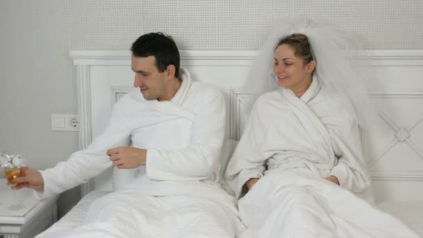 Vtipná vdaná dvojice novomanželů v hotelu leží na posteli a pije šampaňské ze svatebních brýlí. Nevěsta v závoji a bílý župan s ženichem baví a radostný polibek - Záběry, video