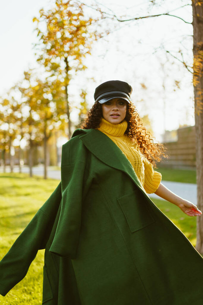 swirling around in the autumn park on the grass girl in a leather cap and bright yellow sweater throwing on her shoulder a green coat - Foto, imagen