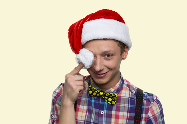 glimlachende tiener jongen is sluiten zijn oog met santa hoeden pompon. - Foto, afbeelding