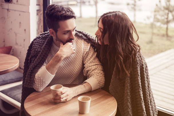 若い夫婦はとても寒かったので、カフェに入った。暖かく保つために抱擁ラグの下に座ってください。話、浮気、話し合いなさい。異性愛者との関係の概念。コーヒー休憩。男は思った. - 写真・画像