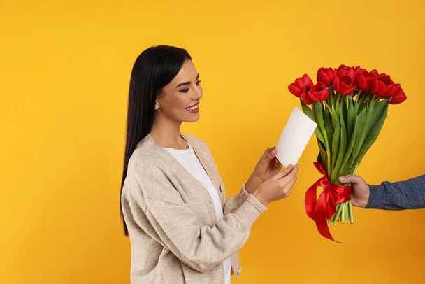 Happy woman receiving red tulip bouquet and greeting card from man on yellow background. 8th of March celebration - Fotografie, Obrázek
