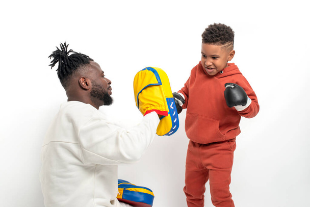 Hombre africano de piel oscura y su pequeño hijo pasan tiempo juntos. Papá enseña a su hijo a boxear contra fondo blanco - Foto, Imagen