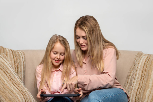 Egy hosszú hajú, rózsaszín inget és farmert viselő szőke anya és lánya, akik otthon ülnek a kanapén egy laptoppal..   - Fotó, kép