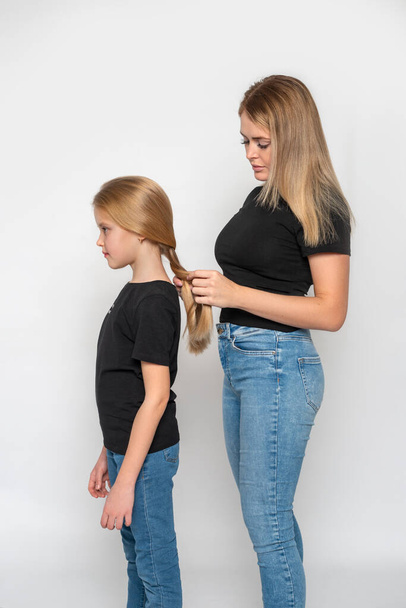 ママは娘のためにピグテールを編む彼らは同じ黒いシャツとジーンズを着ている - 写真・画像