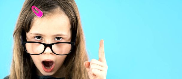 Lähikuva muotokuva lapsi koulun tyttö yllään etsivät lasit nosta syyttävällä sormella vuonna Minulla on idea ele eristetty sinisellä pohjalla. - Valokuva, kuva
