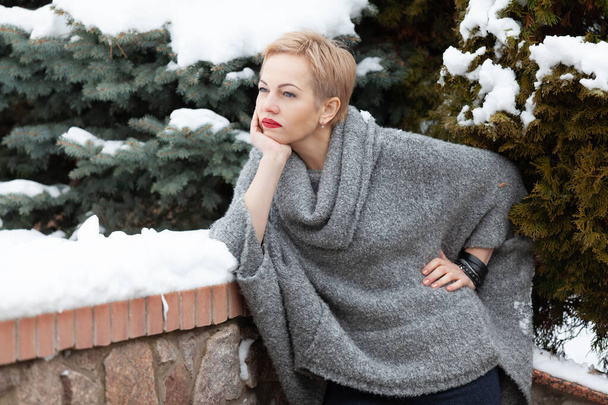 beautiful blonde in a warm sweater is standing in a winter yard in front of a green fir and leaning on a stone fenc - Фото, изображение