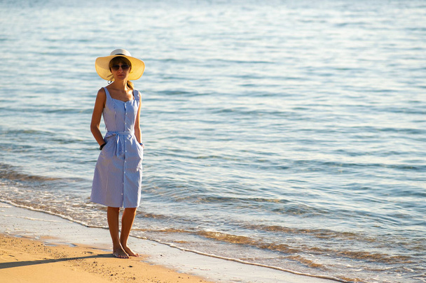Young woman in straw hat and a dress walking alone on empty sand beach at sea shore. Lonely tourist girl looking at horizon over calm ocean surface on vacation trip. - Photo, Image