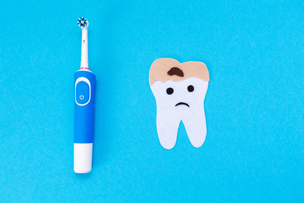 An electric toothbrush and a tooth cut out of felt, affected by caries. Blue background. Flat lay. The concept of modern dental products. - Photo, Image