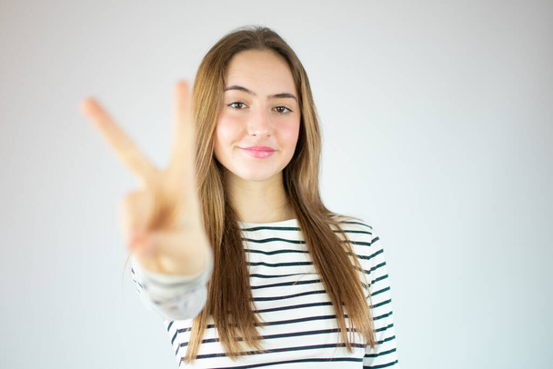 Portret van een lachend meisje met overwinningsteken en kijkend naar camera geïsoleerd over witte achtergrond - Foto, afbeelding