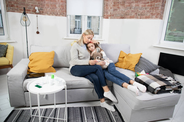 Young attractive Caucasian woman mother, laying on the soft gray sofa at home, hugging her sick teen daughters and checking her temperature, looking at the thermometer after measuring - 写真・画像