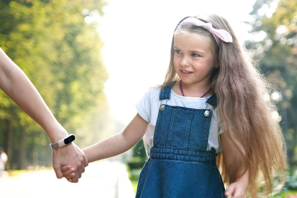 Fiatal anya és kislánya hosszú hajjal, kéz a kézben sétálnak a nyári parkban.. - Fotó, kép