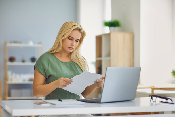 Serious young woman sitting at desk at home reading official notification letter - Фото, изображение