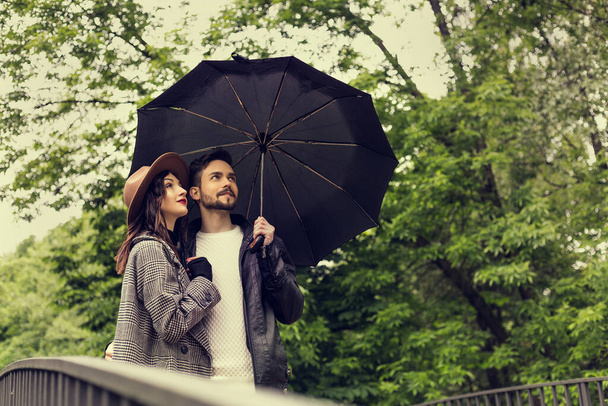 Portret heteroseksualnej pary stojącej na zewnątrz na moście w parku miejskim przy rzece w pochmurną deszczową pogodę z czarnym parasolem. Obraz z miejsca kopiowania i bokeh tła. - Zdjęcie, obraz