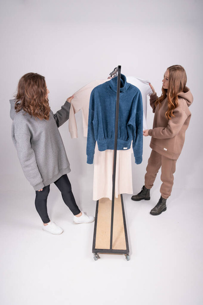 Hermosas amigas haciendo compras eligiendo ropa. Dos mujeres jóvenes navegando a través de un estante de ropa en un estudio, fondo gris claro. Foto de alta calidad - Foto, imagen