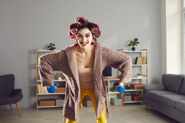 Happy young woman in skincare face mask and hair curlers enjoying sports workout at home - Foto, afbeelding