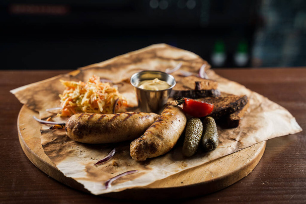 Grilled sausages with beer and vegetables with pickled cucumber, cabbage salad sauce and bread on parchment, top view. - Photo, Image