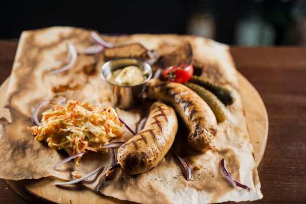 Grilled sausages with beer and vegetables with pickled cucumber, cabbage salad sauce and bread on parchment, top view. - Photo, Image