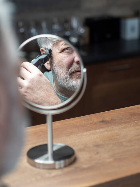 Ein älterer kaukasischer Mann blickt in den Tischspiegel und schneidet sich seine grauen Haare mit einem Elektroschneider. Selbstbeschneidung zu Hause. Kopierraum. - Foto, Bild