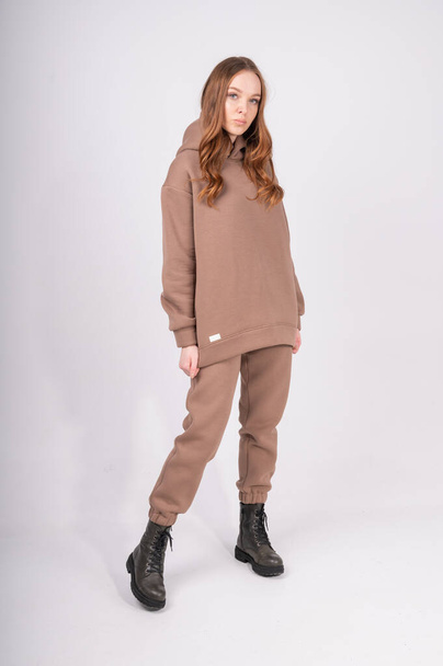 Retrato de comprimento total de mulher jovem em taupe hoodie e elegantes botas grosseiras isoladas em branco. Camisola feminina bege e calças com etiqueta em branco. Conceito de roupa de rua. Foto de alta qualidade - Foto, Imagem