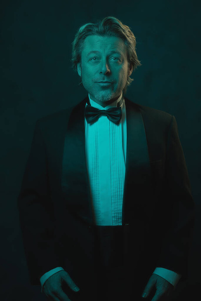 Blonde man wearing tuxedo and bow tie. Lit by blue and orange light against dark wall. - Photo, Image