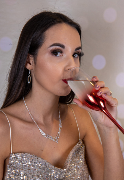 A sexy elegant woman wearing a silver glittery party dress with long brown hair and red lipstick looking like she is ready for an elegant Christmas party or wedding with a glass of wine in her hands - Zdjęcie, obraz