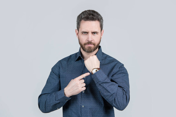 Unshaven επιχειρηματίας σε casual πουκάμισο δείχνουν δάχτυλο στο χέρι ρολόι γκρι φόντο, ώρα εργασίας. - Φωτογραφία, εικόνα