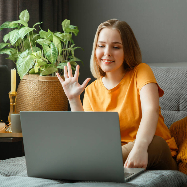 Happy young beautiful woman greets, talking by video call using laptop while sitting on sofa in living room. Remote work study, communication with family and friends in home interior - Foto, Imagem