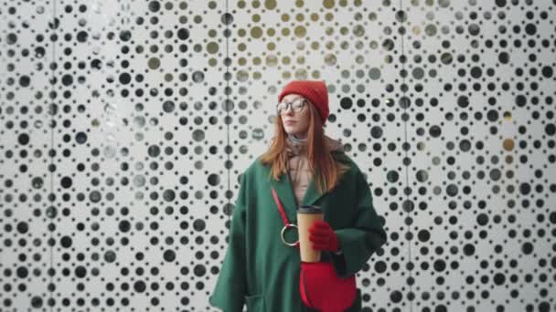 Zoom in shot of young attractive woman in stylish outfit holding disposable coffee cup and posing for camera while standing outdoors in front of modern urban building on winter day - Footage, Video