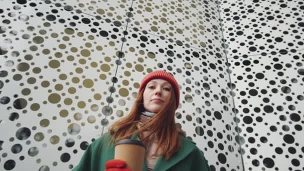Niskokątne ujęcie portretowe młodej pięknej kobiety w odzieży wierzchniej, trzymającej filiżankę kawy i patrzącej w kamerę stojącej na zewnątrz przed nowoczesnym budynkiem miejskim z perforowaną ścianą - Materiał filmowy, wideo