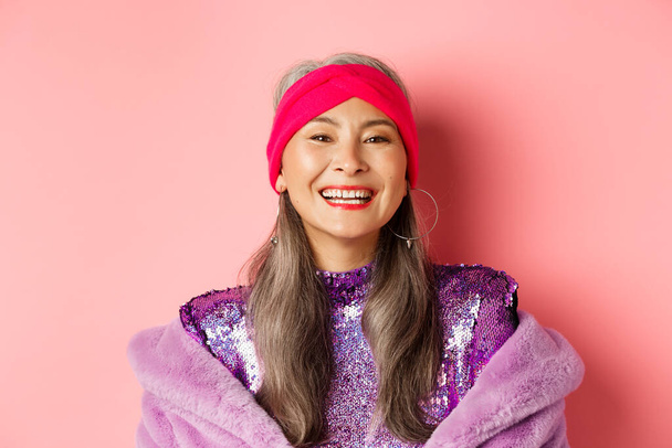 Detailní záběr šťastné asijské starší dáma v módní fialové oblečení, směje se a usmívá se na kameru, baví na růžovém pozadí - Fotografie, Obrázek