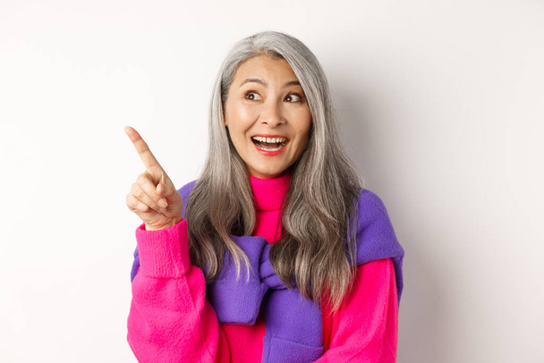 Gros plan de la coréenne élégante femme âgée avec les cheveux gris regardant et pointant coin supérieur gauche, montrant l'offre de promotion, debout sur fond blanc - Photo, image