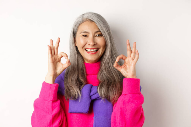 Primer plano de mujer asiática satisfecha con el pelo gris, sonriendo complacido y mostrando signos OK, aprobar y como producto, alabando algo impresionante, fondo blanco - Foto, imagen
