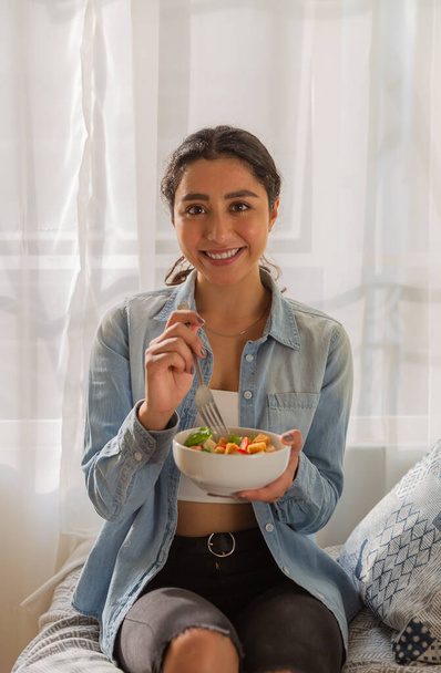 Young brunette woman is smiling and eating a salad. She is sitting on a sofa in her room - Photo, Image