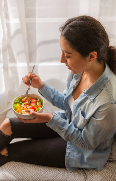 Jeune femme brune sourit et mange une salade. Elle est assise sur un canapé dans sa chambre - Photo, image