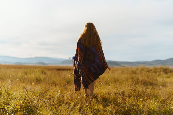 on the steppe grass in the distance towards the hills goes barefoot girl in a long cape with long red hair - Photo, Image