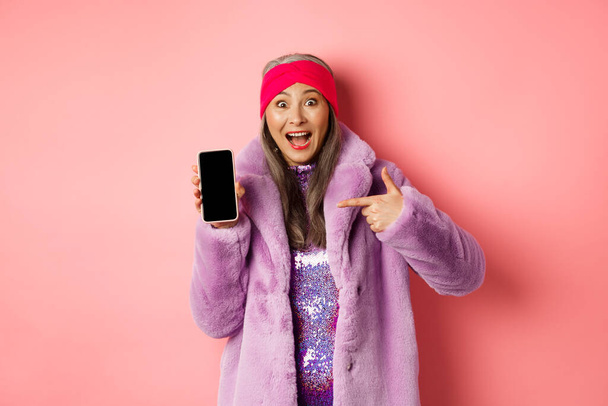 Online shopping and fashion concept. Stylish asian senior woman in faux fur coat pointing finger at blank smartphone screen, smiling happy, showing application on phone - Photo, Image