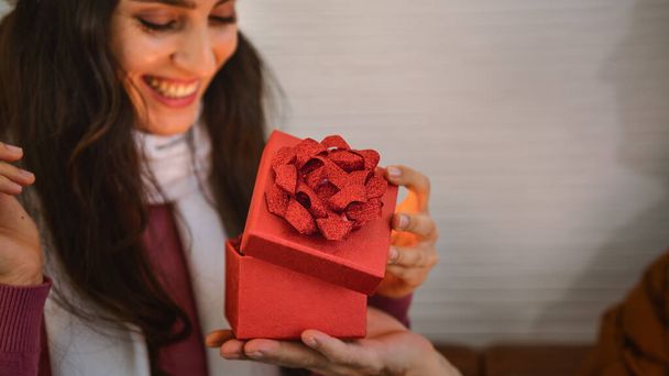 close up of gift in red box on hand of man giving gift to woman with surprised smiling face for anneversary celebration. - Foto, afbeelding