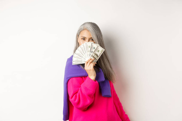 Nákupní koncept. Bohatá a módní korejská starší žena skrývající tvář za penězi, dívající se na kameru, stojící nad bílým pozadím - Fotografie, Obrázek