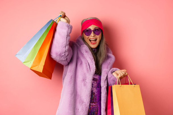 Κομψό ασιατική ηλικιωμένη γυναίκα πηγαίνει για ψώνια, φορώντας μοντέρνα ρούχα και γυαλιά ηλίου, κρατώντας τσάντες κατάστημα και χορό ευτυχισμένη, ροζ φόντο - Φωτογραφία, εικόνα